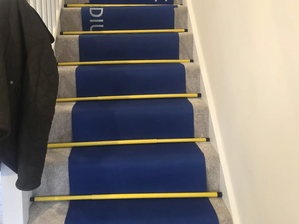 Blue stair runner held down with yellow rods on a carpeted staircase